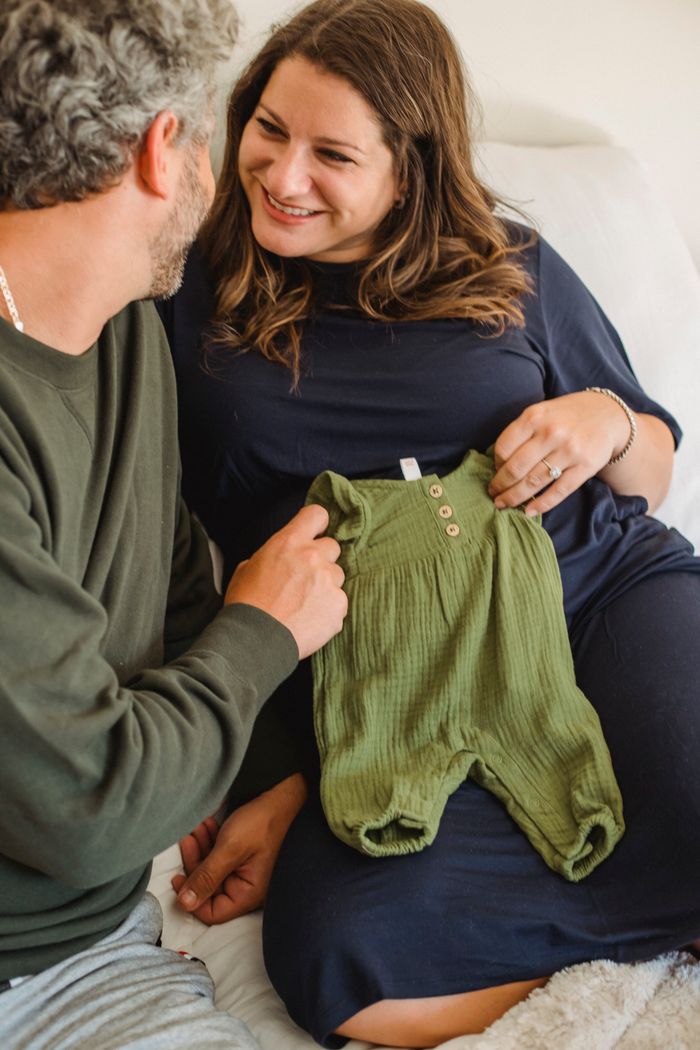 Crop romantic pregnant couple relaxing on couch while preparing apparel for infant baby and looking at each other