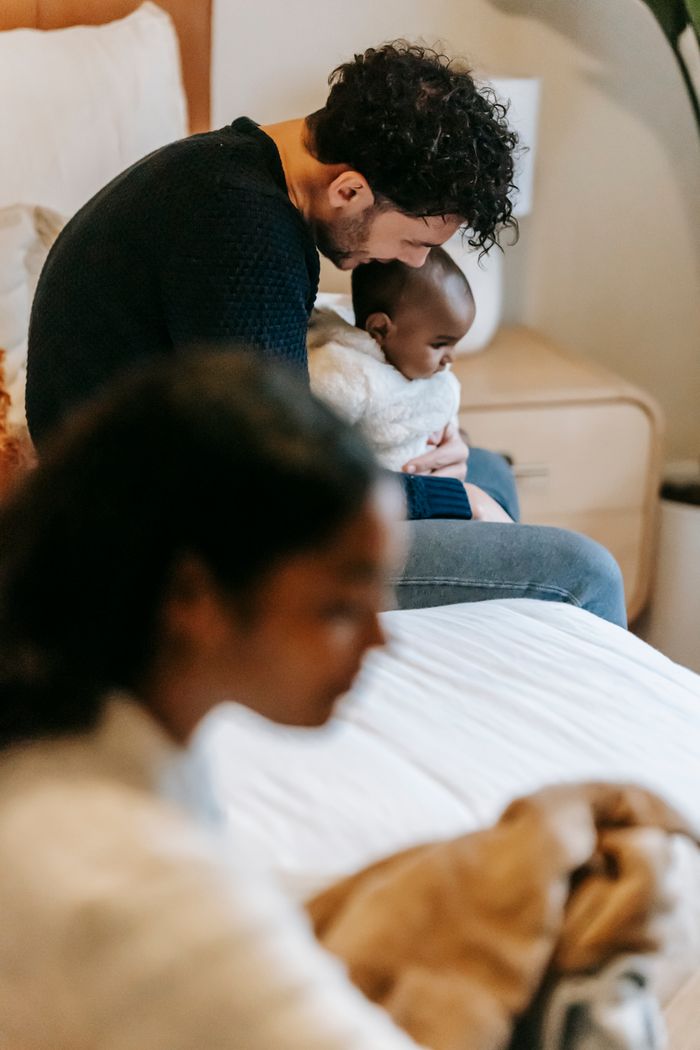 Side view of young ethnic father hugging cute black baby while sitting on comfortable bed near wife at home